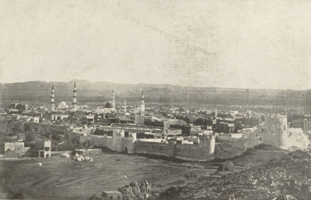 Black and white panorama of Medina about 1880: city walls in the foreground, the Prophet's Mosque with its dome and minarets behind, and mountains on the horizon