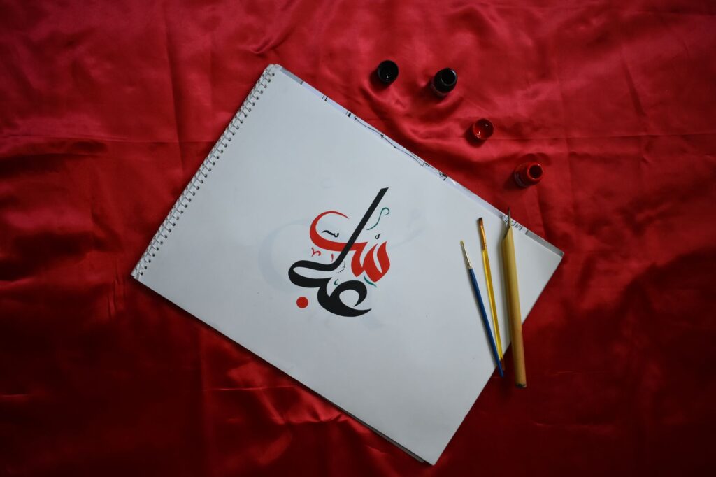 A top-down view of Arabic calligraphy on paper, styled text, and pens on red fabric, an image for Written by Humans? How We Know Muhammad Did Not Author the Quran