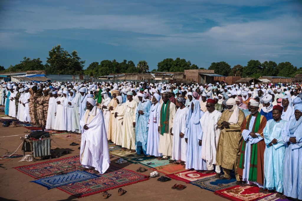 Large gathering of Muslims praying outdoors in traditional attire.