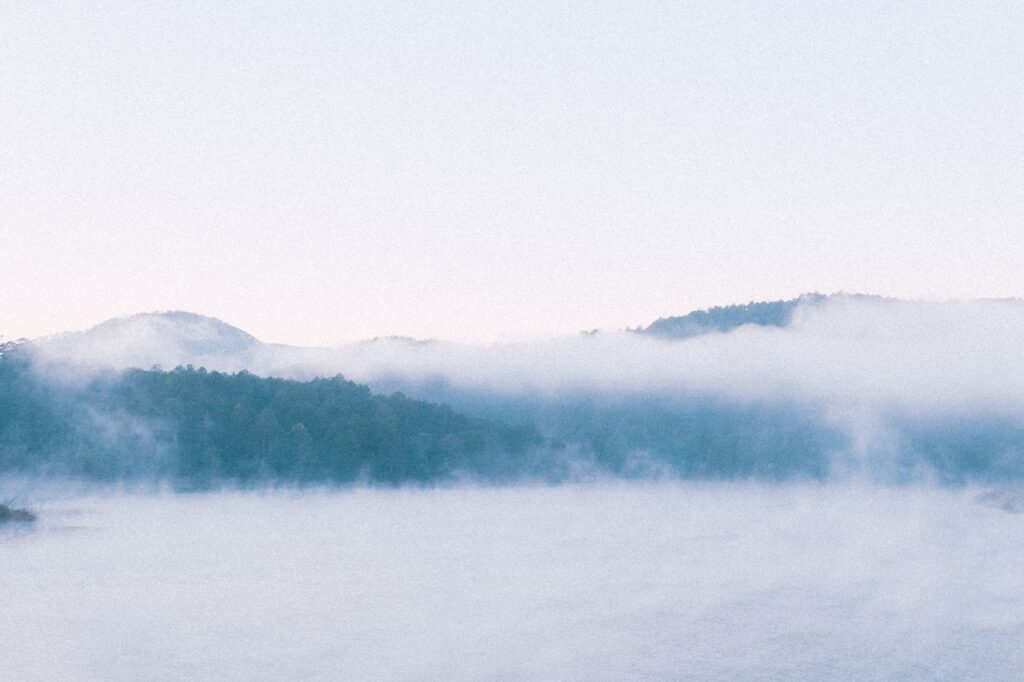 Peaceful foggy lake scene with mist-covered trees and hills, evoking tranquility and calm.