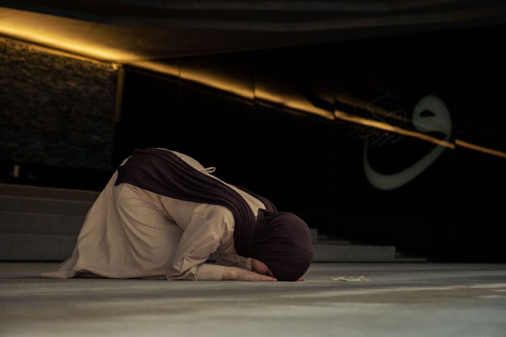 A woman in hijab is kneeling and praying inside a mosque, creating a serene religious atmosphere.