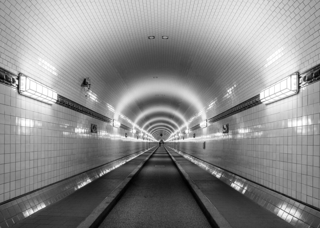 Black and white photo of a symmetrical underground tunnel in Hamburg, featuring tiled walls and diminishing perspective.