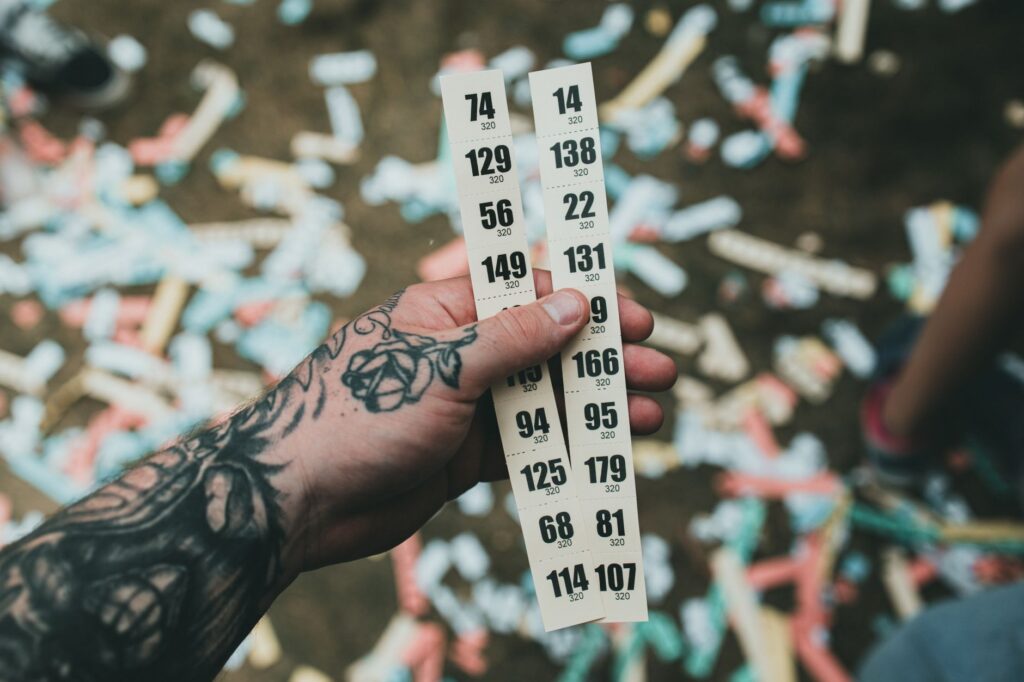 Close-up of a tattooed hand holding raffle tickets amidst confetti at a festival setting, an image for God Rewards Faith and Punishes Doubt: Just a Loyalty Test
