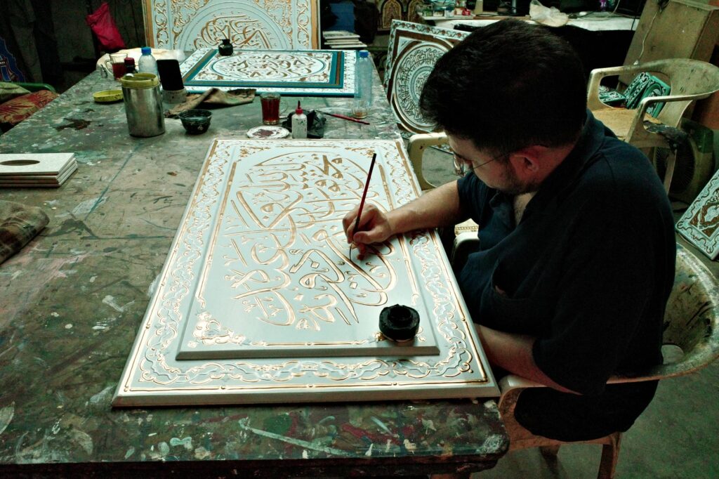 Artisan meticulously painting traditional Islamic calligraphy on a decorative panel indoors.