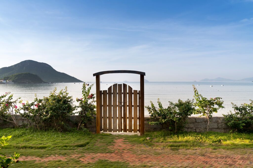 Stone fence with green bushes and grass along and wooden gate to seashore with picturesque view on calm sea with mountain on horizon and blue sky in sunny…