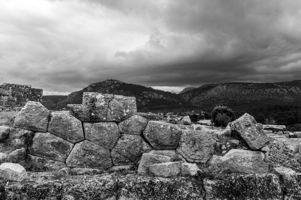 Dramatic black and white photo of stone ruins against a mountainous backdrop under cloudy skies.