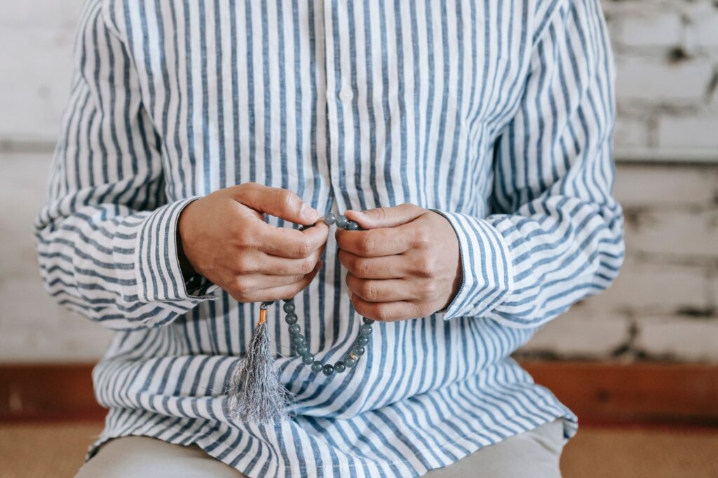 A person wearing a striped shirt holds prayer beads close, symbolizing faith and meditation, an image for Practising Without Belief: What Ritual Means When Faith Is Gone