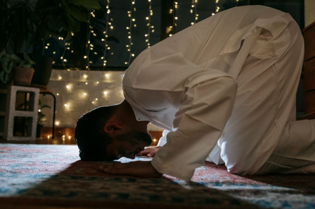 A man in traditional attire bows in prayer on a rug lit by string lights, symbolizing faith and devotion.