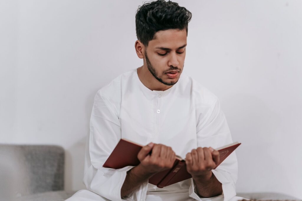 A young man in Islamic attire intimately reading the Quran indoors, an image for First Time Reading the Quran: What Happens