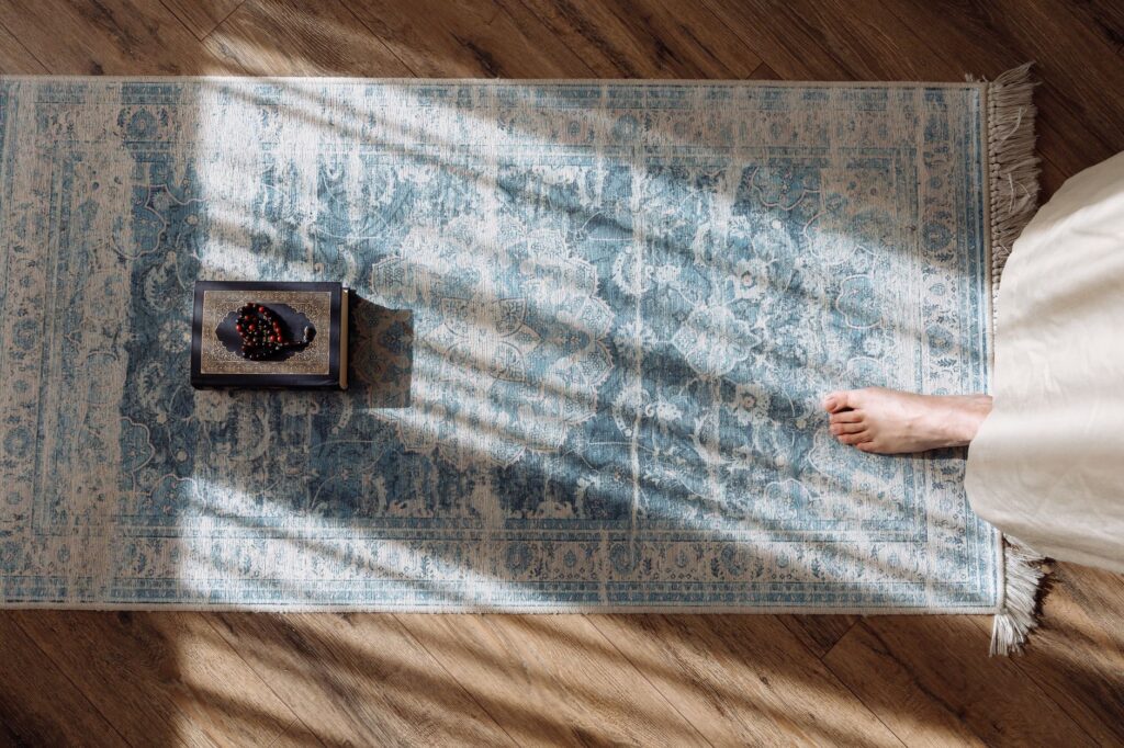 Top view of a prayer setup with Koran and beads on a patterned rug, sunlight casting shadows, an image for Does God Need Our Prayers